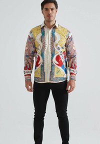 Homme portant une chemise à manches longues colorée avec des motifs de soleil et abstraits, assortie à un pantalon noir, se tenant devant un fond clair uni.