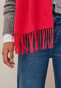 Écharpe rouge vif aux bords frangés, en tissu doux. Portée sur un pull gris et associée à un jean en denim bleu.