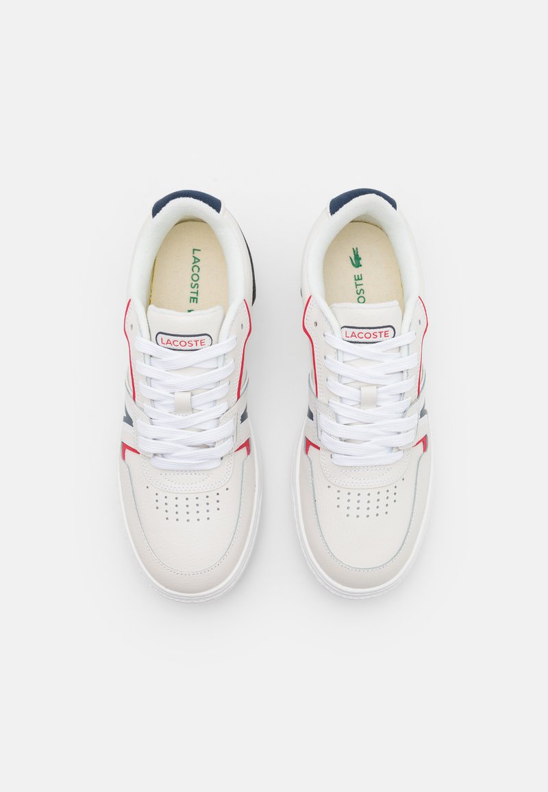 Baskets en cuir blanc avec des accents rouges et bleus marine, présentant une perforation à l'avant, des lacets blancs plats et un logo vert sur la semelle intérieure.