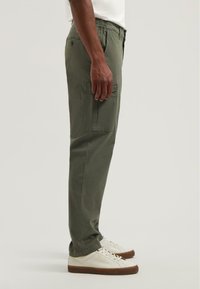 Olijfgroene cargobroek met zijzakken, elastische tailleband en taps toelopende pijpen. In combinatie met witte sneakers en een gestructureerde stof.