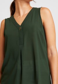 Top verde sin mangas con escote en V, que presenta un detalle de un solo botón dorado, textura suave y un ajuste relajado con un pequeño pliegue frontal.