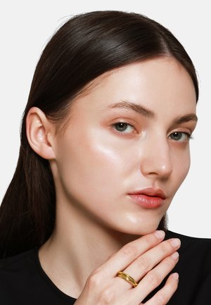 Goldring am Finger, glatte Haut mit natürlichem Glanz, glattes braunes Haar, minimales Make-up und neutrale Kleidung vor einem schlichten Hintergrund.
