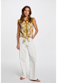 Mosterdkleurige mouwloze blouse met een knoopsluiting aan de voorkant, ingewikkelde witte paisleypatronen en een geknoopt zoom. Gepaard met witte wijde broek.