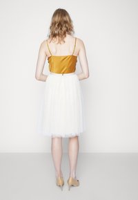 Nainen seisoo kasvot seinää kohti, pukeutuneena sinapinkeltaiseen olkaimelliseen toppiin, valkoiseen polvenmittaiseen hameeseen ja beigekorollisiin kenkiin.