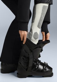 Černá lyžařská bota s nastavitelnými přezkami, obutá na šedé a bílé vzorované ponožce. Strukturovaný materiál s matným povrchem.