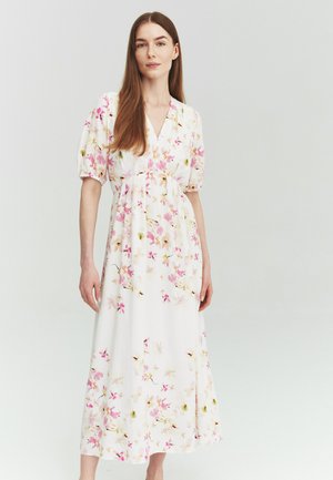 Donna con lunghi capelli castani che indossa un vestito midi bianco con motivo floreale e maniche ampie, in piedi davanti a uno sfondo chiaro e uniforme.