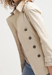 Trench-coat beige avec un design à double boutonnage, des boutons noirs et un col cranté. Le matériau est lisse, avec des poches latérales et une coupe cintrée.