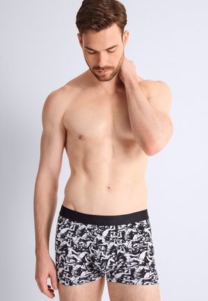 Homme torse nu regardant vers le bas, portant un boxer noir et blanc à motifs, une main reposant sur son cou sur un fond clair.