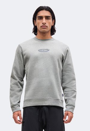 Uomo con capelli scuri di media lunghezza indossa una felpa Patagonia grigio chiaro e pantaloni neri, in piedi davanti a uno sfondo chiaro uniforme.
