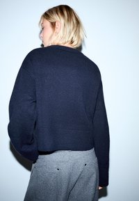 Marinblå stickad tröja med en kortad design och ribbad nederkant. Bärs med grå strukturerade byxor, sett bakifrån.