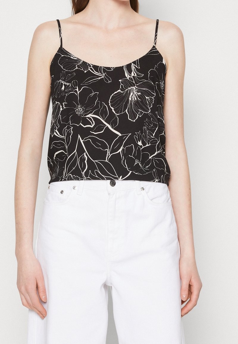 Femme portant un caraco noir à imprimé floral blanc et fines bretelles, associé à un pantalon blanc taille haute sur un fond uni.