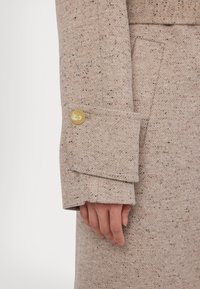 Bežový textúrovaný kabát s jemnými tmavými škvrnami, so klasickým strihom, prehnutým manžetom a zlatým gombíkom na rukáve.