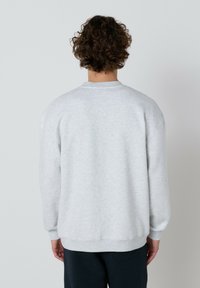 Muž s kučeravými vlasmi, oblečený v svetlosivom mikině a tmavých nohaviciach, stojí tvárou k čistému bielému pozadiu.