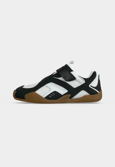Basket profil bas avec tige en mesh blanc, superpositions noires, semelle en caoutchouc marron et une bande Velcro noire sur le dessus.