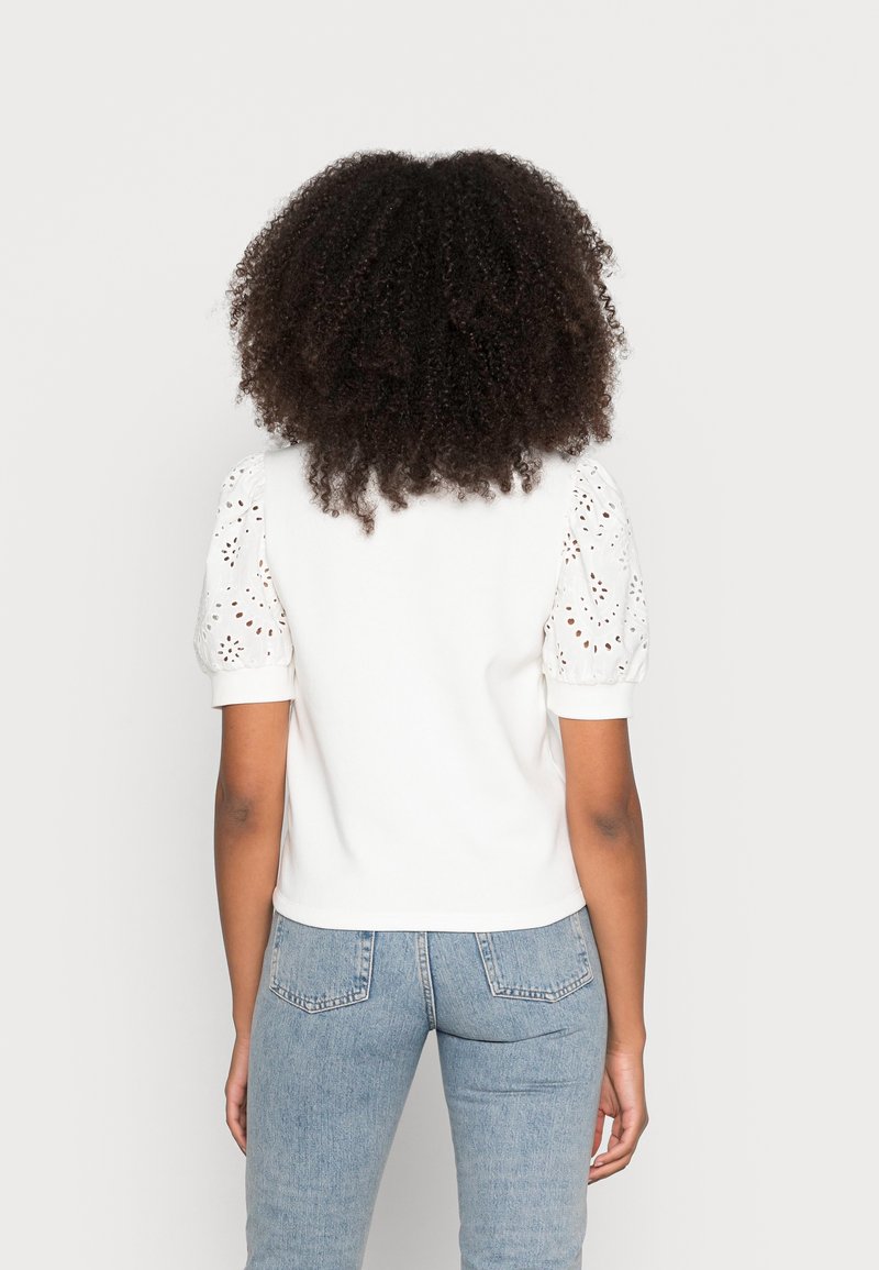 Woman with curly hair wearing a white top with eyelet puff sleeves and light blue denim jeans, shown from the back on a plain background.