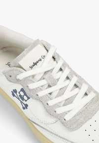 Baskets en suède blanc et gris avec des lacets blancs, un logo tête de mort et os croisés sur le côté, et une semelle beige texturée.