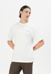 White cotton t-shirt with short sleeves, round neckline, and a chest pocket featuring blue text reading "The Regal." Smooth texture, casual fit.