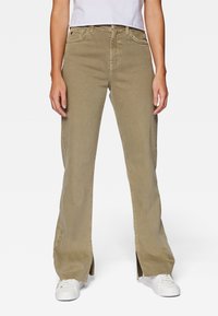 Donna che indossa pantaloni beige a gamba larga con fessure frontali, abbinati a sneakers bianche e una t-shirt bianca infilata dentro.