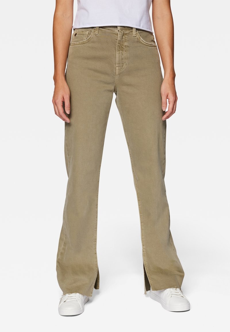 Donna che indossa pantaloni beige a gamba larga con fessure frontali, abbinati a sneakers bianche e una t-shirt bianca infilata dentro.
