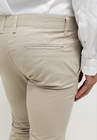 Béžové bavlněné kalhoty mají štíhlý střih, decentní detail zadní kapsy a hladkou texturu. V pozadí je vidět bílá košile.