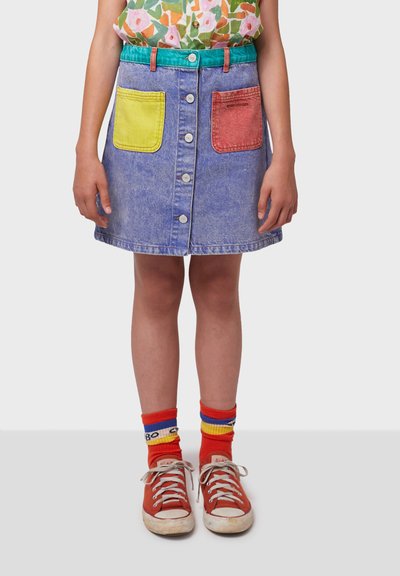 Jupe en denim avec une forme évasée, dotée de poches avant jaunes et rouges, de boutons en argent et d'une taille turquoise. Assortie à des baskets rouges.