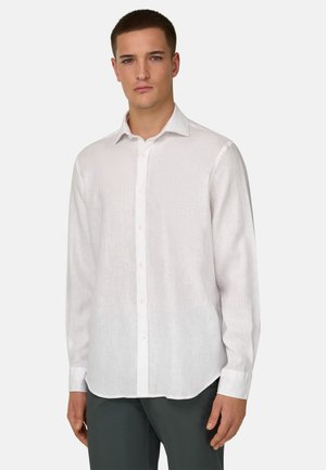 Jeune homme portant une chemise blanche à manches longues boutonnée et un pantalon foncé, debout devant un fond clair uni.