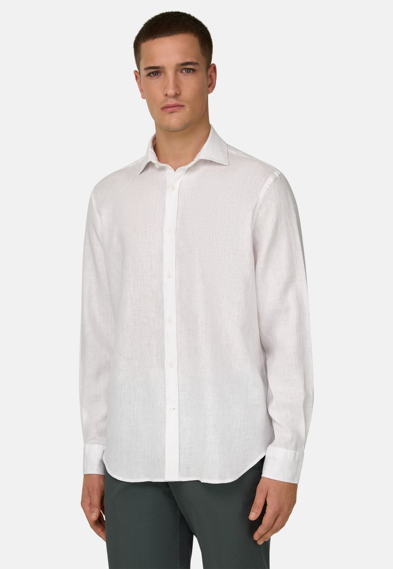 Jeune homme portant une chemise blanche à manches longues boutonnée et un pantalon foncé, debout devant un fond clair uni.
