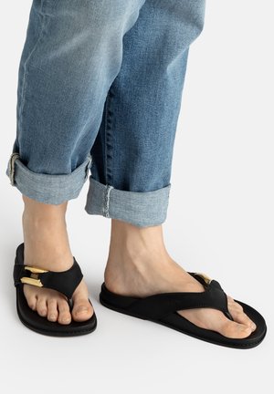 Füße mit schwarzen Zehensandalen mit goldenen Schnallendetails, kombiniert mit blauen hochgekrempelten Jeans auf weißem Hintergrund.