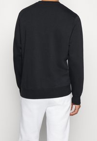 Man i en enkel svart långärmad sweatshirt och vita byxor, sedd bakifrån mot en neutral bakgrund.