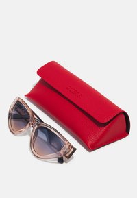 Occhiali da sole a forma di occhio di gatto rosa con lenti sfumate, accanto a un elegante astuccio in pelle rossa con texture, caratterizzato da un logo impresso.