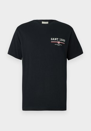 Schwarzes T-Shirt mit kurzen Ärmeln, auf dem "GANT 1949" in weißer und roter Schrift sowie einem kleinen Wappenlogo auf der oberen linken Brust abgebildet sind.