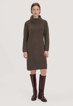 Frau steht und trägt ein braunes, langärmliges Strickkleid mit Rollkragen und knielange dunkelbraune Lederstiefel vor einem einfarbigen Hintergrund.