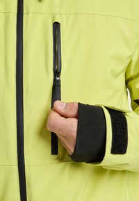 Heldere gele waterdichte jas met een close-up van een ritszak en een zwart elastisch manchet met klittenbandsluiting. Soepele textuur.