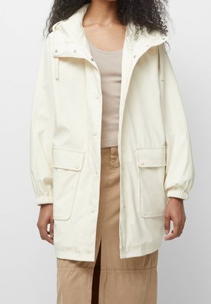 Femme portant un imperméable à capuche blanc cassé avec boutons-pression et grandes poches avant sur une jupe beige et un débardeur.