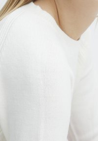 Gros plan sur une personne portant un pull en maille blanc, montrant l'épaule et une partie du cou avec une peau claire et des cheveux blonds.