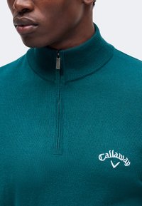 Gros plan d'une personne portant un pull zippé bleu sarcelle avec un col montant et un logo Callaway blanc sur la poitrine.