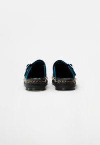 Zöldes-kék velúr slip-on cipők fekete gumitalppal, állítható pántokkal és sárga varrással a széleken. Hátsó nézet látható.