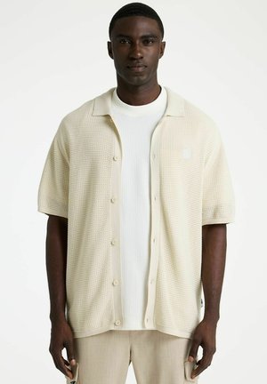Mann trägt beiges, kurzärmliges, geknöpftes Strickhemd über weißem, strukturiertem Oberteil und beigen Hosen, steht vor schlichtem weißem Hintergrund.