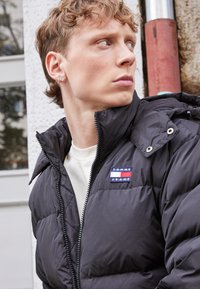 Giacca a vento nera con chiusura a zip, colletto prominente e logo ricamato "TOMMY JEANS". Indossata sopra una camicia di colore chiaro.