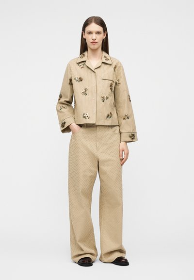 Γυναίκα που στέκεται φορώντας μπεζ ριγέ σακάκι με λουλουδάτη κεντητική και φαρδιές μπεζ παντελόνες με διακριτικό πουά σχέδιο και μαύρα παπούτσια.