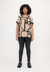 Top beige a maniche corte con motivi geometrici astratti neri, abbinato a leggings neri lucidi e scarpe con zeppa nere decorate con borchie.
