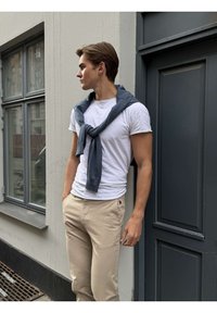 Mladý muž v béžových nohaviciach a bielom tričku so svetlomodrým svetrom prehodeným cez plecia, opierajúci sa o sivé dvere, pozerá doľava.