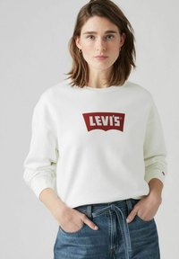 Felpa bianca con un grande logo rosso di Levi's. Maniche lunghe, colletto a giro e vestibilità rilassata. Abbinata a jeans in denim blu a vita alta.