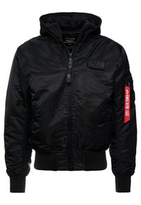 Giubbotto bomber nero con zip frontale, polsini e orlo a costine, e cappuccio rimovibile. Presenta una toppa con logo e un'etichetta rossa "Rimuovere prima del volo".