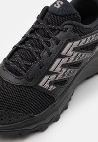 Černá sportovní bota s prodyšným svrškem, texturovanými detaily a gumovou podrážkou. Obsahuje reflexní prvky a kontrastní systém šněrování.