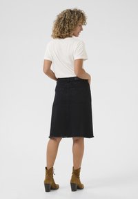 Jupe en denim noir en forme de A avec un ourlet effiloché, deux poches arrière, associée à une chemise blanche à manches courtes et des bottines en daim marron.