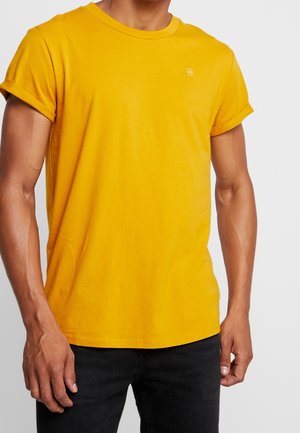 Homme portant un t-shirt jaune uni à manches courtes avec revers roulés et un pantalon noir devant un fond gris clair.
