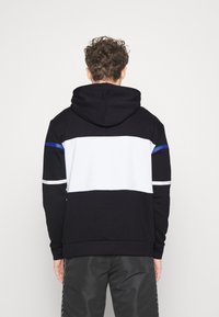 Mikina s kapucí v černé, bílé a modré barvě. Obsahuje design s blokováním barev a horizontální pruhy. Vyrobena z měkké tkaniny, má dlouhé rukávy a volný střih.