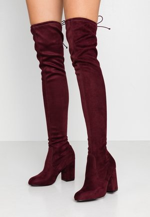 Femme portant des bottes en daim bordeaux au-dessus du genou avec talons carrés et liens à l'arrière sur un sol blanc.