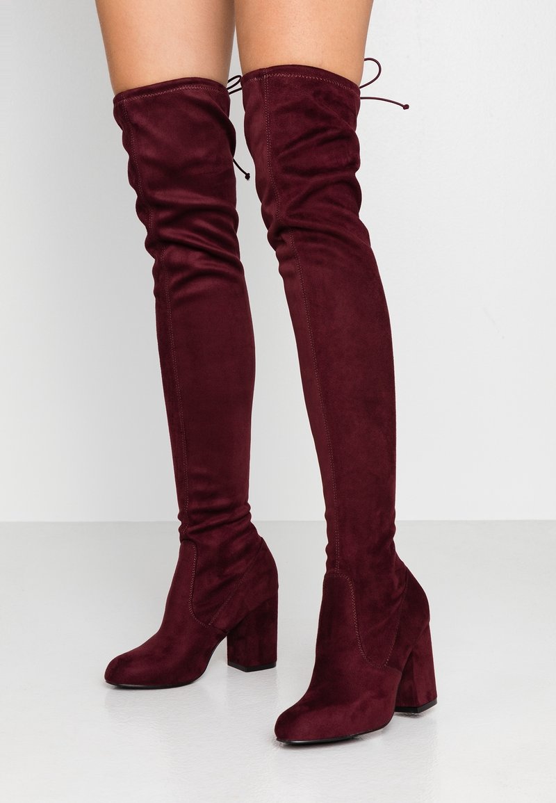 Femme portant des bottes en daim bordeaux au-dessus du genou avec talons carrés et liens à l'arrière sur un sol blanc.